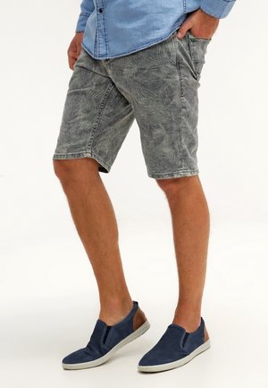 Grå shorts med et subtilt palmebladsmønster, lavet af materiale, der ligner denim, parret med marineblå slip-on sko med tan accenter.