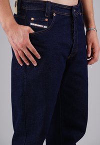 Dunkelblaue Jeans aus Denim mit kupfernen Nieten, orangefarbenen Nähten und einer Vordertasche mit einem kleinen dekorativen Etikett. Entspannter Schnitt.