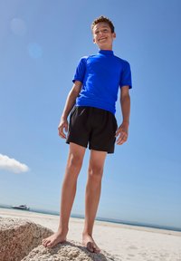 Oseba, ki stoji bosa na skali na plaži, nosi modro kratkodlako rash guard majico in črne kopalne hlače, v ozadju je jasno nebo.