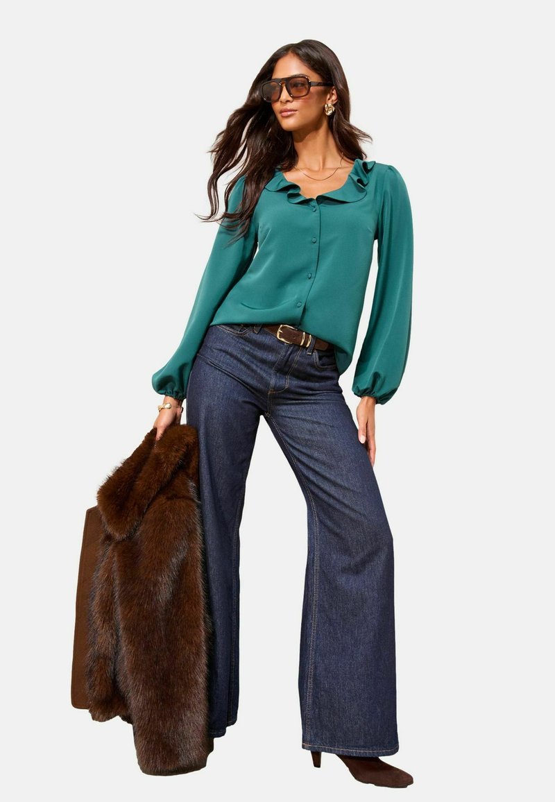 Teal blouse with ruffled collar, brown faux fur coat, dark blue flared jeans, and brown ankle boots. Model wears oversized sunglasses.