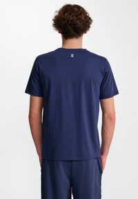 Mörkblå bomullströja med rund hals och korta ärmar, med ett litet vitt emblem på baksidan av halsen. Slät textur, avslappnad passform.