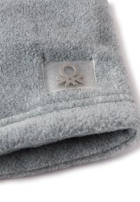 Scarf in pile grigio con una texture morbida, caratterizzato da un'etichetta chiara con un simbolo in argento. Dettaglio del bordo ripiegato visibile su un lato.