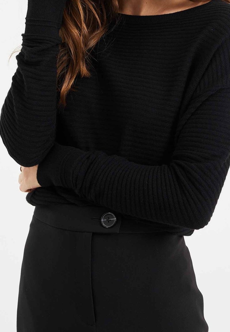 Femme portant un pull noir côtelé à manches longues, rentré dans un pantalon noir taille haute avec une fermeture à un seul bouton.