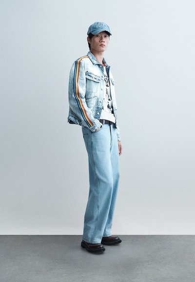 Νεαρός άνδρας που φοράει τζιν μπουφάν με ρίγες ουράνιου τόξου, ανοιχτόχρωμα μπλε τζιν, γραφικό μπλουζάκι, μπλε καπέλο και σκούρα παπούτσια στέκεται μπροστά σε μια απλή φόντο.