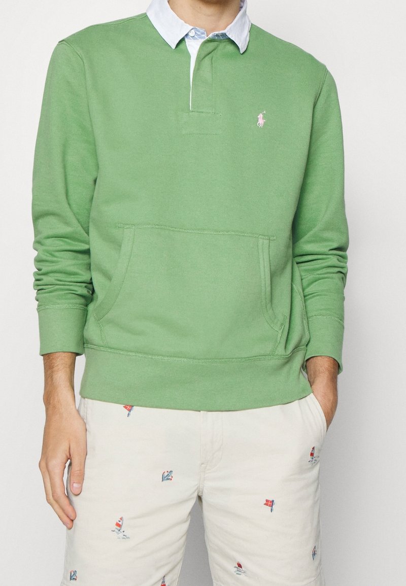 Uomo che indossa una polo a maniche lunghe verde chiaro con colletto bianco e pantaloni beige ricamati con piccole barche a vela colorate.