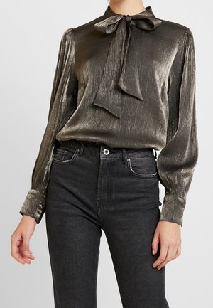 Kobieta ubrana w metaliczną, brązową, długorękawową bluzkę z wiązaną kokardą przy kołnierzu oraz czarne, wysokowysokie jeansy na jednolitym tle.