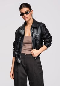 Femme portant une veste en cuir noire, un haut marron sans manches, un pantalon large noir et des lunettes de soleil ovales noires, sur un fond uni.