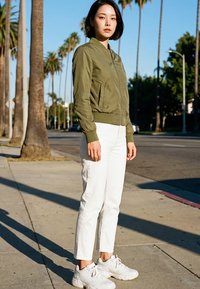 Fiatal nő áll a járdán, olíva zöld dzsekit, fehér nadrágot és fehér tornacipőt visel, mögötte pálmafák és utca látható.