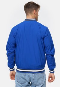 Mand med ryggen til iført en blå bomberjakke med hvid- og sortstribede manchetter og krave, parret med lyseblå denimjeans.