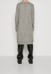 Dlouhý šedý pletený kardigan s žebrovanými manžetami a lemem, s volným střihem a bočními rozparky, v kombinaci s lesklými černými kalhotami s širokými nohavicemi.