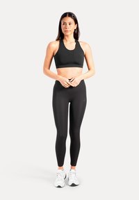 Zwarte sportbeha met een brede band, gecombineerd met zwarte leggings met hoge taille. Model draagt witte sneakers; minimalistisch ontwerp en gladde textuur.