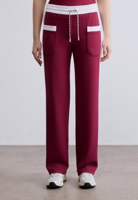 Μπορντό φόρμα με λευκό κορδόνι και πλαϊνές τσέπες, με άνετη εφαρμογή και ομαλή υφή. Λευκές ρίγες ως διακοσμητικό στοιχείο στις τσέπες.