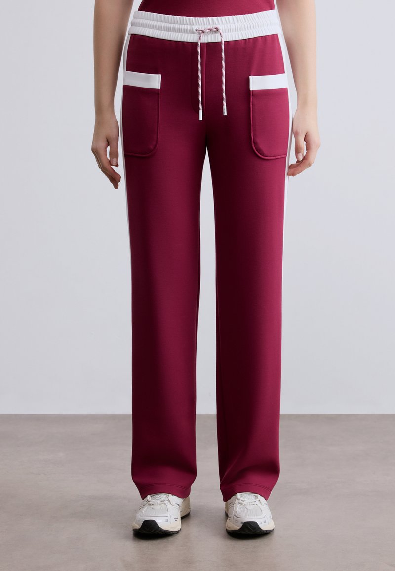 Μπορντό φόρμα με λευκό κορδόνι και πλαϊνές τσέπες, με άνετη εφαρμογή και ομαλή υφή. Λευκές ρίγες ως διακοσμητικό στοιχείο στις τσέπες.