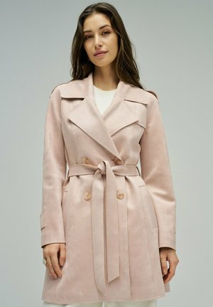 Mujer con un abrigo trench rosa claro con cinturón, solapas anchas y botones de doble pecho, de pie frente a un fondo gris neutro.