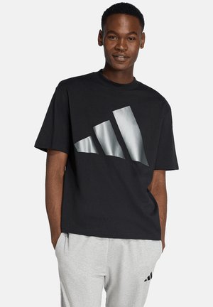 Uomo che indossa una maglietta nera con un grande logo Adidas argentato e pantaloni della tuta Adidas grigio chiaro con le mani nelle tasche.