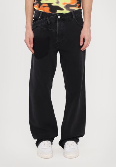 Jeans larghi in denim nero scuro con una tasca a toppa distintiva sulla coscia sinistra, caratterizzati da un classico design a cinque tasche e passanti per la cintura.