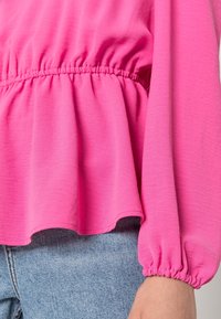 Blusa rosa con vita arricciata e maniche lunghe e gonfie. Il tessuto è leggero e leggermente testurizzato. L'orlo è curvo e più lungo sul retro.
