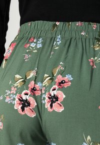 Calças florais com uma cintura elástica, apresentando um tecido verde adornado com flores rosas e azuis, criando um padrão texturizado.