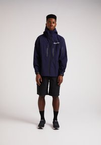 Marineblå vandtæt jakke med høj krave, sorte lynlåslommer og justerbare manchetter, parret med sorte cargo-shorts og sorte sneakers.