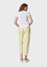 Top bianco a maniche corte abbinato a pantaloni giallo chiaro, che mostrano un design aderente e una texture liscia. Le calzature presentano accenti dorati.