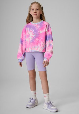 Młoda dziewczyna ubrana w różowo-fioletową bluzę tie-dye, lawendowe krótkie spodenki rowerowe, białe skarpetki i lawendowe trampki stoi na szarym tle.