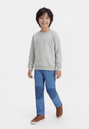 Fant z temnimi lasmi, oblečen v sivo majico z dolgimi rokavi, modre hlače z blazinicami na kolenih in rjave superge, stoji in se smeji na belem ozadju.