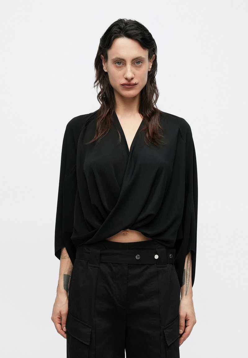 Fekete, laza szabású felső V-nyakkivágással és redőzött kialakítással, magasderekú fekete cargo nadrággal párosítva, oldalzsebekkel és patent díszítésekkel.