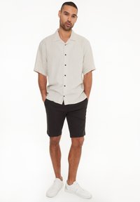 Helles beige, kurzärmliges Hemd mit Knöpfen in Schwarz, kombiniert mit dunklen Shorts und weißen Sneakers. Lässiger, lockerer Schnitt.