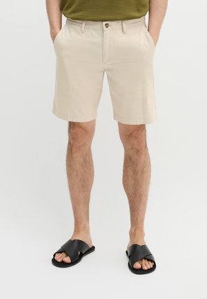 Man die beige korte broek en zwarte slides draagt, handen in de zakken, staande tegen een eenvoudige lichte achtergrond.