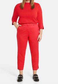 Pull en maille rouge à manches trois-quarts associé à un pantalon rouge ajusté avec des fentes latérales. Chaussures noires ornées d'accents métalliques.