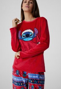 Czerwona koszulka z długim rękawem z nadrukiem niebieskiej postaci z kreskówki w czapce Mikołaja. Dopasowane czerwone spodnie piżamowe z świątecznym wzorem z postacią Stitche'a i dodatkami.