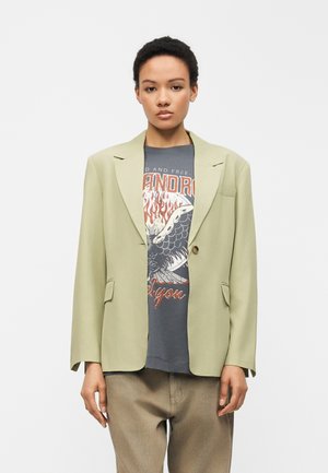 Jeune personne aux cheveux courts et bouclés, portant un blazer vert clair sur un t-shirt gris foncé graphique et un pantalon beige, debout devant un fond blanc.