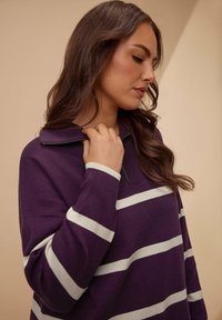 Lila Fleece-Pullover mit weißen horizontalen Streifen, ausgestattet mit einem Reißverschlusskragen und lässiger Passform. Texturiertes Gewebe mit leichtem Glanz.