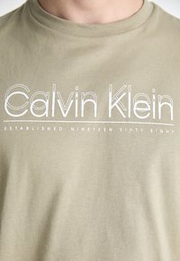 Olivově zelené bavlněné tričko s bílým logem "Calvin Klein" a textem "ZALOŽENO V DEVATENÁCTI ŠESTI OSMDESÁTI" níže. Klasický kulatý výstřih.