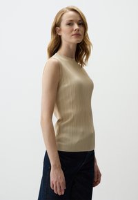 Top senza maniche beige a coste con scollatura alta, design aderente e tessuto texturizzato. Abbinato a jeans scuri per un contrasto.