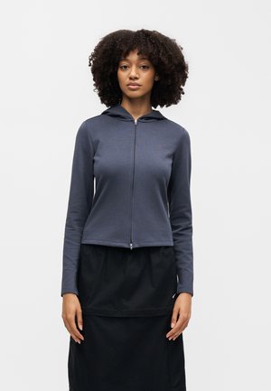 Vrouw met krullend haar draagt een marineblauwe hoodie met rits en een zwarte rok, staande tegen een effen lichte achtergrond, naar voren gericht.