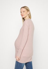Sweater de maternidade em rosa claro, de mangas compridas, em tecido suave, apresentando um corte descontraído e detalhes em ribana nos punhos e na barra, combinado com jeans azuis.