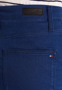 Blå denimjeans med en svart läderlapp vid midjan. Har en bakficka med en liten röd, vit och blå logotyp. Slät struktur.