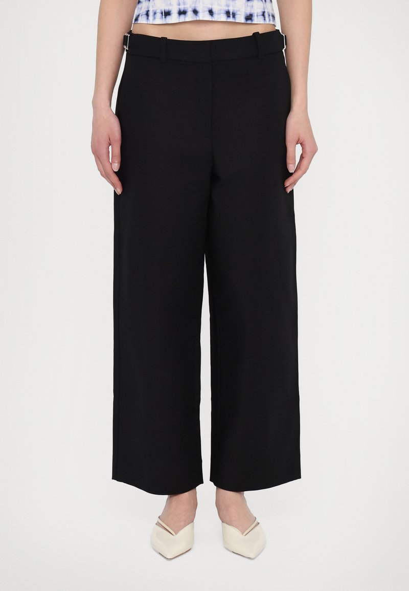 Black wide-leg trousers with a smooth fabric finish, featuring a flat front, belt loops, and side pockets, paired with cream pointed shoes.