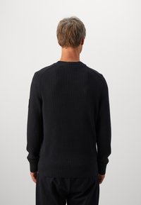 Man with short hair wearing a black ribbed sweater, shown from the back against a plain light background.