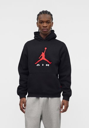 Junge Person mit geflochtenem Haar, die einen schwarzen Hoodie mit rotem Basketball-Logo und graue Adidas-Sweatpants trägt, stehend vor einem einfarbigen Hintergrund.