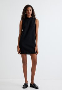 Schwarzes ärmelloses Kleid mit hohem Kragen und geradem Schnitt; verfügt über dekorative Knöpfe an der Schulter. Getragen mit schwarzen Slippern.