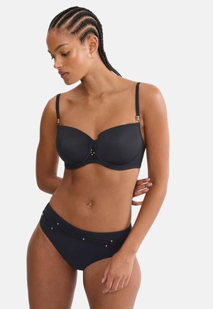 Woman with braided hair wearing a black bikini with gold bead accents, standing with one arm behind her back, looking down to the left.