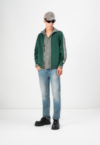 Veste zippée verte avec des rayures blanches, chemise à carreaux en dessous, jeans bleu clair et chaussures noires épaisses. Une casquette et des lunettes de soleil complètent le look.
