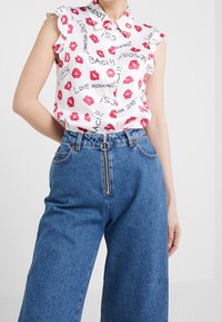 Vit blus med röda läpptryck och svart text, har volangärmar. Kombineras med vida blå denimjeans med dragkedjedetalj.