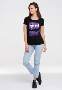 Black t-shirt featuring a purple graphic with a cartoon character and "Woodstock '69" text. Paired with light blue jeans and black sneakers.