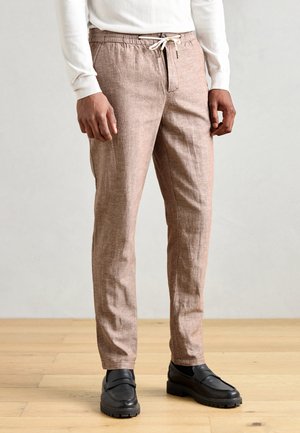 Pantalones de mezcla de lino de color marrón claro con cintura ajustable, bolsillos laterales y corte slim. Combinados con mocasines de cuero negros.