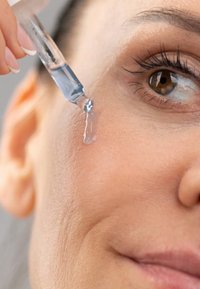 Przezroczysta pipeta uwalniająca przezroczysty niebieski serum na gładką, jasną skórę. Zbliżenie podkreśla delikatne rzęsy oraz szczegółową strukturę skóry.