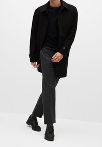 Cappotto di lana nero con chiusura a bottoni, camicia scura a maniche lunghe, pantaloni grigi con trama, e scarpe sportive nere. Abito casual dal design moderno.