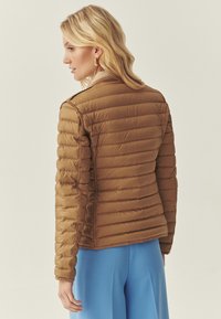 Femme aux cheveux blonds portant une veste matelassée marron et un pantalon bleu, vue de profil sur un fond neutre.